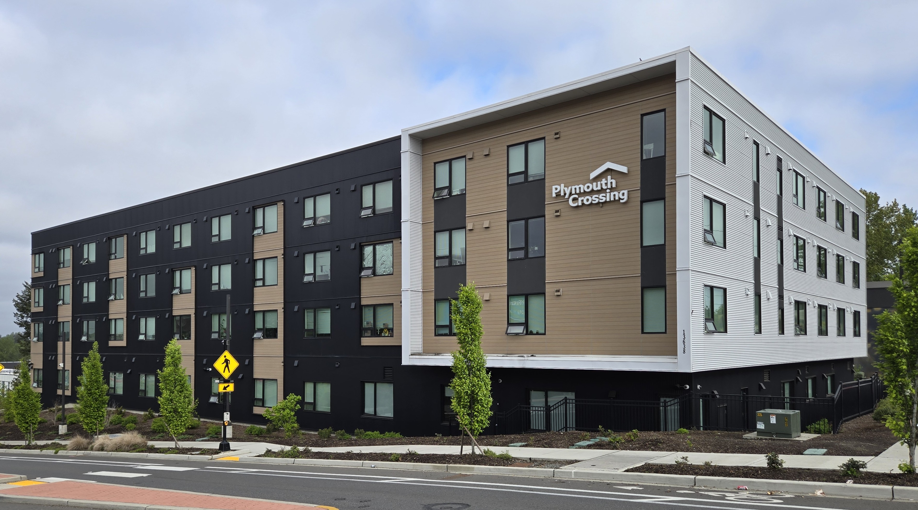The image shows a modern multi-story building with a combination of black and beige exterior walls, featuring large windows and a flat roofline. There is signage at the top that reads Plymouth Crossing.In front of the building, there are trees and a sidewalk leading to a crosswalk. The sky is overcast, suggesting an urban environment.